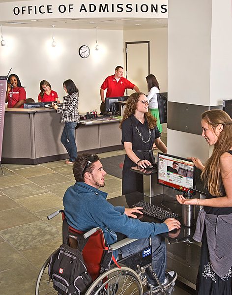 Student in wheelchair receives computer help while other students ask questions at the Admissions counter.