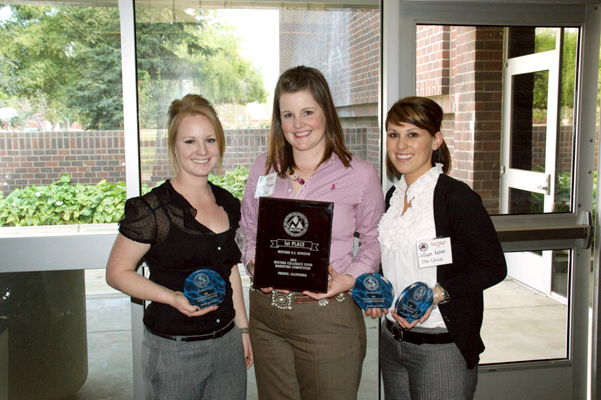 Three women pose smiling while standing in front of a large window.  They are dressed in business-casual attire, slacks and button-down blouses with big white name tags. All three hold one or more award plaques. The woman in the center holds the largest, 1st place award plaque.