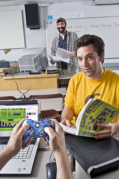 Man in yellow shirt holds book on game design while man wearing hat in background smiles.