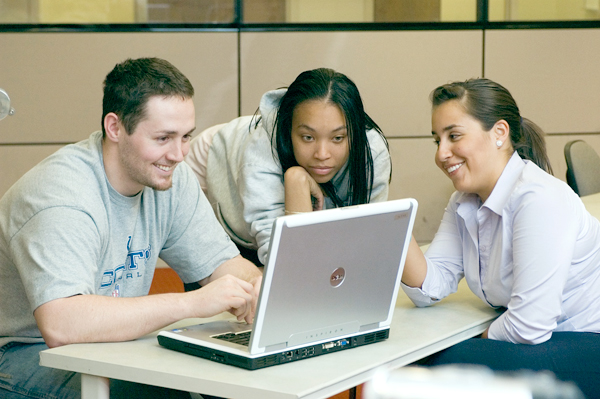 Three people huddle around a laptop computer at the end of a long, narrow table which cascades out of the screen to the right.  Left: A smiling man wearing a t-shirt, sits in front of the laptop typing.  Center: A woman in a hooded sweatshirt leans onto the table looking at the laptop monitor resting her cheek on her up-folded arm.  Across the table, sits a woman in business attire, who smiles while pointing at the monitor.