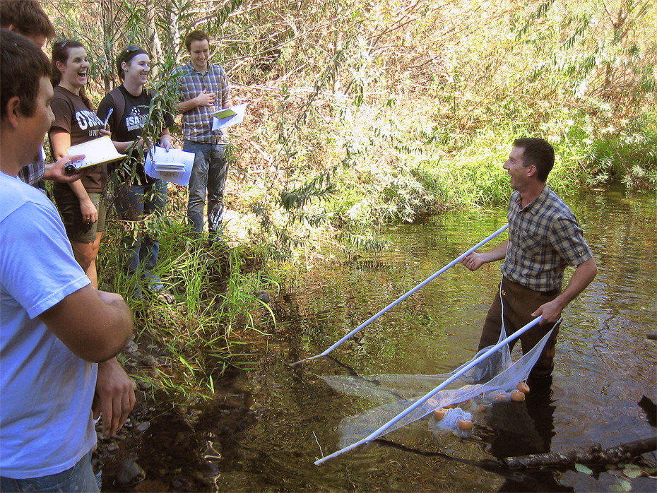 Man in plaid shirt stands in water holding a net by two metal bars with both hands. He shares a laugh with several students who stand on the shore (to the left) holding notebooks while facing the man in the water.