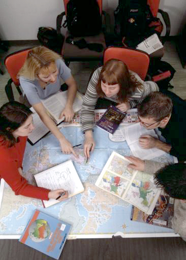 Five students sit around a table covered by a large world map. Two students point to a location on the map while the other two students look on and write notes.