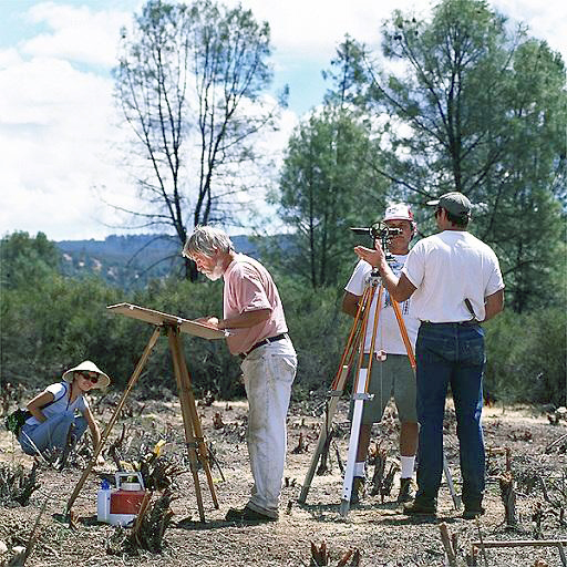 Three men stand around several surveying instruments. One man, center, wearing a salmon-colored shirt tucked into white pants leans over a board on top of a tri-pod, touching it with his left hand.  A woman, right, wearing sunglasses and a conical hat kneels in the background
