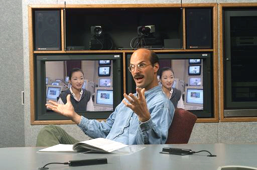 A man faces left wearing glasses sits at a table gesturing with his hands apart, palms up, fingers spread open, speaking. Behind him in a audio/visual cabinet are speakers encasing two free-standing video cameras above two monitors each showing the same woman on screen.