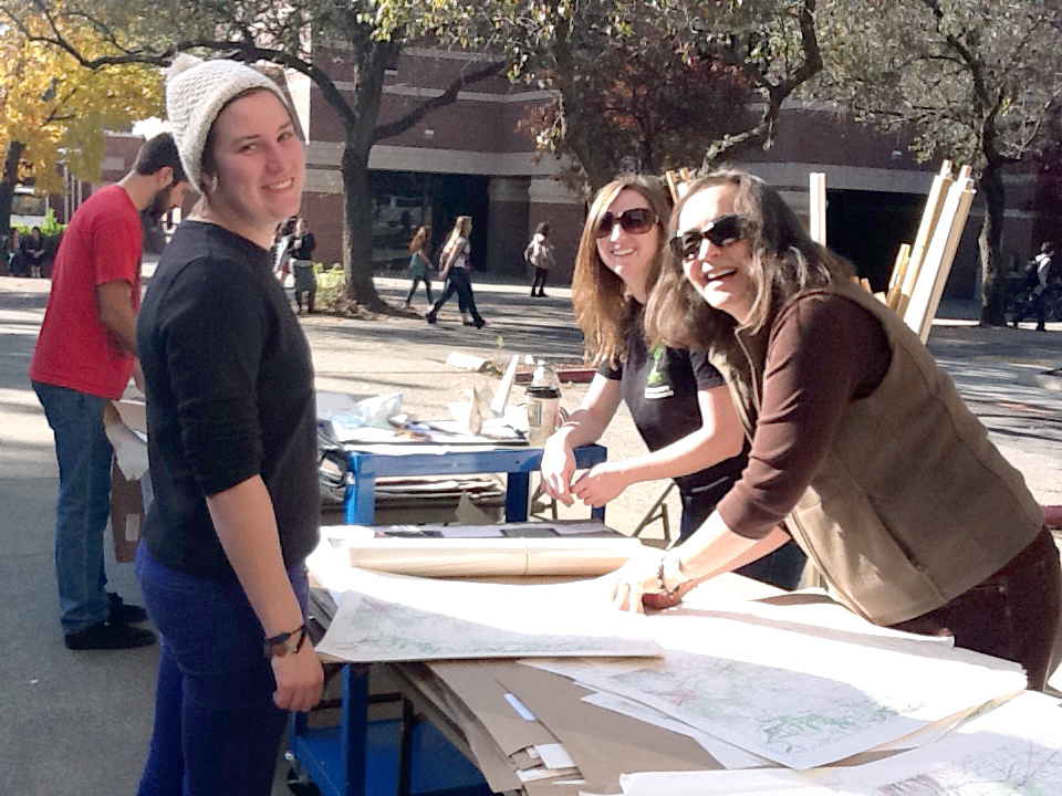 A long table runs from the foreground to the background covered in collections of maps.  One woman, left, wearing a snow cap, bracelet, and dark shirt with jeans stands facing right as two women on the other side of the table lean over it, handling the maps.  All women face and smile to the camera.