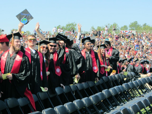 Graduates stand in a line facing left, wearing caps, gowns, sashes, and smiles.  One man, second from left, wears sunglasses and holds his decorated grad cap in the air.