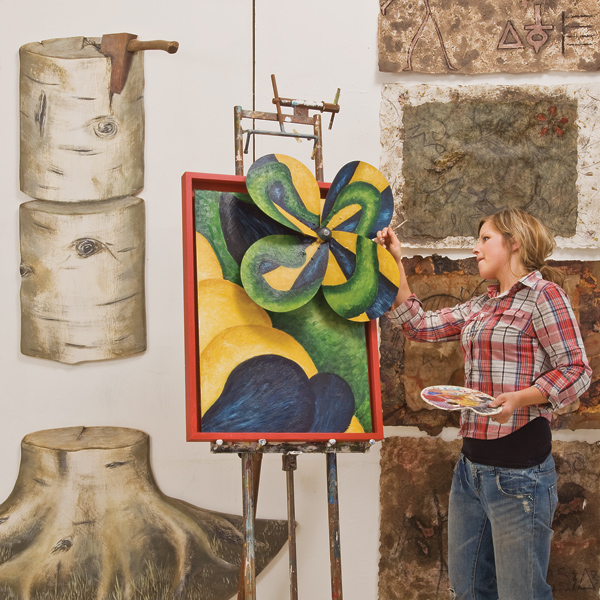 Woman (right) holds paint pallette in left hand, paints pinwheel jutting from framed painting.