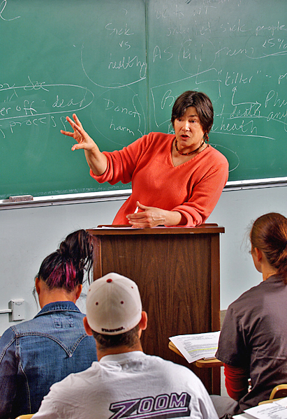 Woman in orange sweater holding chalk in one hand, leans on a podium, and gestures with her other hand, fingers spreading, as she talks.