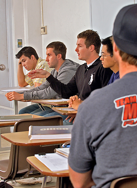 Male students sitting in a row of desks against the wall, listen as one row member talks. 