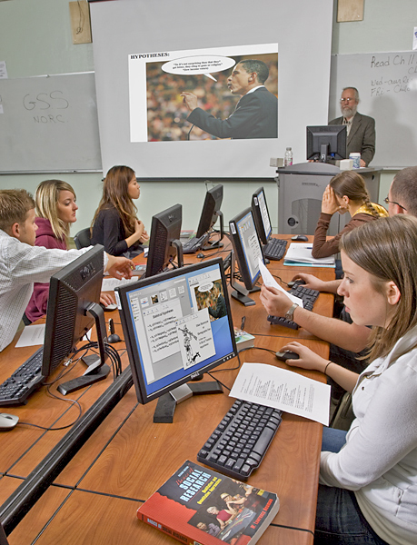 Screen in front of computer lab class displays a photo of Barack Obama with a speech bubble and the caption "Hypothesis." Student computers have same photo on monitors as students walk-through a presentation.