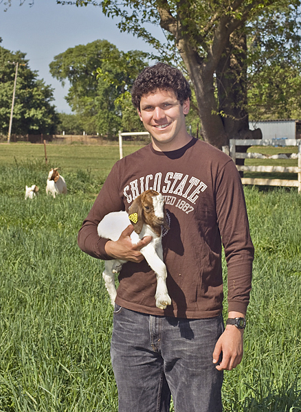 Smiling man wearing a brown long-sleeved Chico State shirt holds a kid (a young goat) in his right arm, while two more goats look on in the background.
