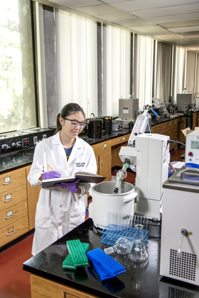 One female student, wearing a lab coat and gloves, holds a book while looking into a container. She stands behind a black topped table with a graduated cylinder, test tube racks, and machines on it.