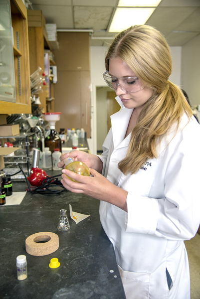 Woman, wearing a lab coat and holding a beaker of sorts, examines it closely. She stands at a black-topped counter with numerous bottles and machines.