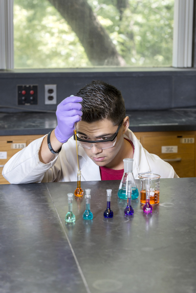 Man wearing glasses, works, kneeling, at lab table holding a straw-like dropper slightly into the top of a resting test tube. The test tube is filled with a transparent orange-tinged liquid.  Also on the table are five other test tubes, each with a different colored transparent liquid (from left to right: green, teal, blue, purple, and pink), a graduated cylinder, and a beaker. 