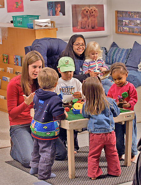 Two adult women interact with several small children. One reads to a girl on her lap, while the other kneels at a table with building blocks where four other children stand and play.