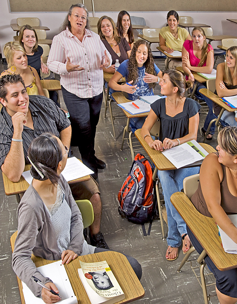 Woman leads classroom discussion while walking through rows of students sitting in desks.