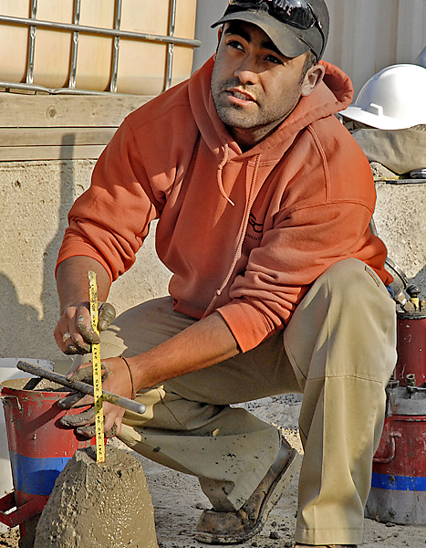 Kneeling man in orange sweatshirt uses tools to determine the height of a conical concrete shape he is working on.
