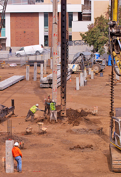 Three construction workers guide a depth measuring device toward the ground next to a foundation support beam during early construction of the Student Services Center.