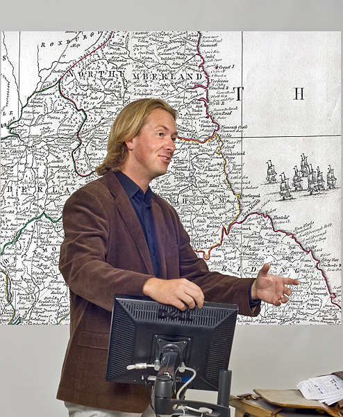 Man in brown blazer stands in front of a map and gestures with hand as he leans on a computer monitor.