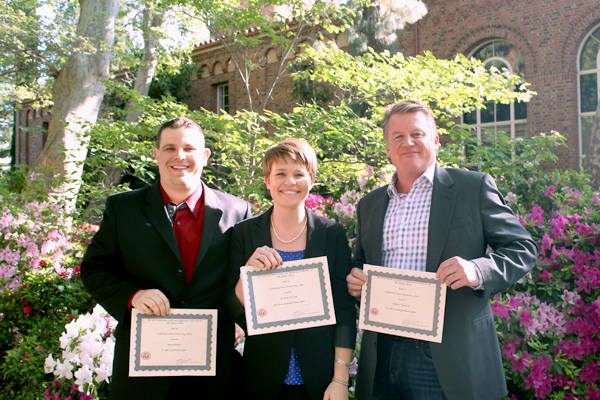 One woman with short hair stands outside flanked by two men on each side.  All three hold winning certificates from the Northern California Regional Conference of Phi Alpha Theta.  The western afternoon sun cascades in from the right of the image over the left shoulders of all three individual.  