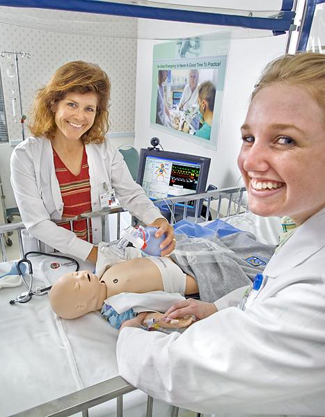 Two women practice maternity ward techniques on a dummy of a "newborn" baby. One holds a manual respirator and the other checks the pulse on the baby's arm with an IV.