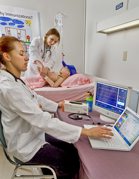 Two women wearing lab coats practice tending to a dummy of a hospitalized young boy. One woman checks the boy's pulse while the other woman interprets information on a couple of digital read-out screens.