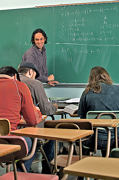 Smirking man leads a discussion at the front of a classroom. He leans on the chalk tray at the board which has several mathematical-type equations written on it.
