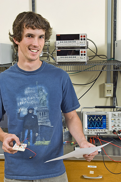 Smiling man holds a piece of machinery in right hand and a sheet of paper in his left. Machine displaying a sin wave pattern with wires hanging out is to his right.