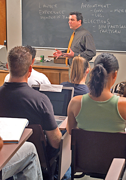 Students sit in classroom listening to a man in a grey shirt and orange tie discuss government and lawyers. 