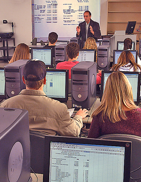 Man stands at front of a computer lab with a screen behind him discussing financial information. Most of the monitors in the room display a spreadsheet of some sort.