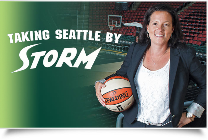 A smiling woman, holding an orange and white basketball on her right hip, stands at half court of the WNBA's Seattle Storm.  She wears a white shirt, with a black jacket over it and black pants.  A rack of five orange and white basketballs rests in front of a green sign in the background.