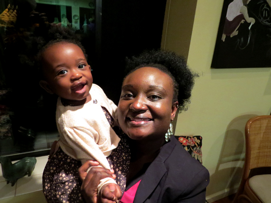 A woman of color holds a baby (suspected to be her child) up near her shoulder.  Both are smiling at the camera.  The baby wears a white sweater over a navy blue dress covered in stars.  The woman wears a black sport coat over a top that has pink in it.