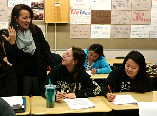 One woman stands with her arm bent towards her, hand near her face, wears a gray scarf and looks down at two other women sitting in desks explaining something.  One seated woman holds a writing utensil and looks up at the woman explaining.  The other seated woman looks and writes on a packet page.  