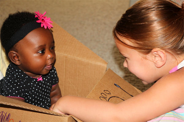 An infant in a box (left) makes faces at another, older, little girl whose face is directly in front of the toddler's face.  The older child reaches inside the box with her left hand and smiles.