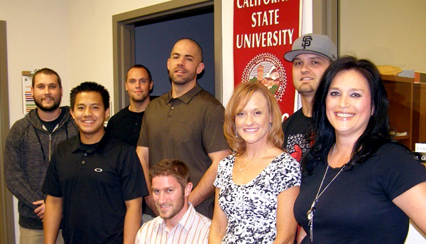 Eight people gather for a photo.  One person sits.  A "California State University, Chico" banner hangs on a wall behind the subjects.