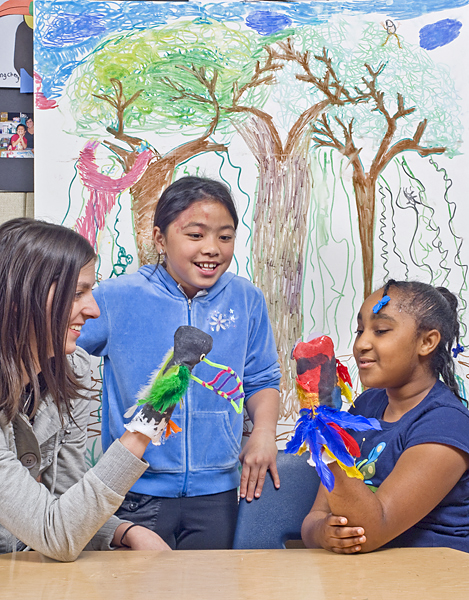 One young girl is entertained by an adult woman and another young girl wearing puppets on their hands.