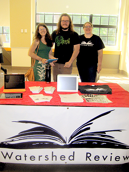 Three people, two women and one man, stand behind a table covered in red with a white banner on the front.  The banner is white with an image of a book laying down (top view) in black and the words "Watershed Review" below it.  The table has brochures, flyers, and a black t-shirt on it.