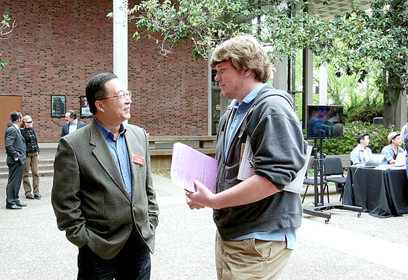 Two men stand on campus in the atrium of the Performing Arts Center (PAC).  Man on the left is older, and wears a brown sports coat with a blue shirt and glasses.  He smiles and looks at the other man (on the right).  Man on right holds a purple sheet of paper in front of him while holding another paper object under his arm.