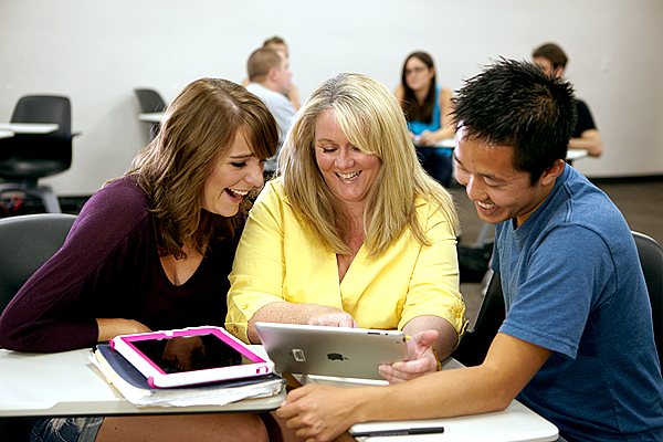 Three people sit at a small table smiling and looking down at a silver tablet.  Woman on left wears a dark purple shirt; woman in middle wears a bright yellow shirt; while man on right wears a lighter blue shirt.  Woman in middle holds tablet in her left hand and points to the screen with her right.