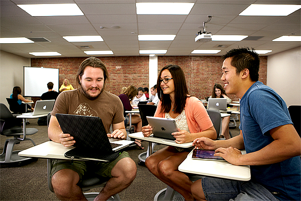 Three students sit in desks and look over at the monitor of a laptop computer held by man on the left, wearing a brown shirt and shorts.  Woman in the middle, holds a tablet wearing a salmon-colored sweater over a white top.  She wears glasses.  Man on right, wears lighter blue shirt and looks towards the left at the monitor.