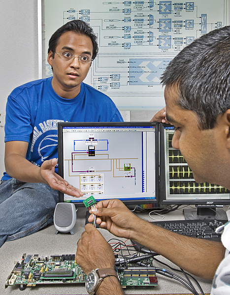 One man sits on a table behind a computer monitor and gestures to another man who is plugging wires into a chip above a motherboard.