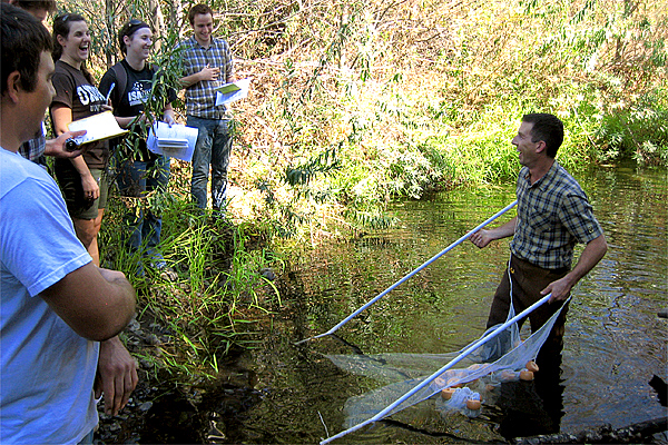 Man in plaid shirt and wading pants smiles big and stands in a creek holding a net with several yellowish objects.  Four to five students stand on the bank watching the man, also smiling, while holding binder paper and a smaller book.