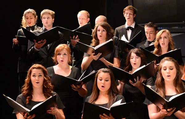 A group of formally dressed people, stand against a blackened stage, under spotlight, holding black binders. All have their mouths opened in the shape of an "o" as if singing a lyric or note with that  sound.