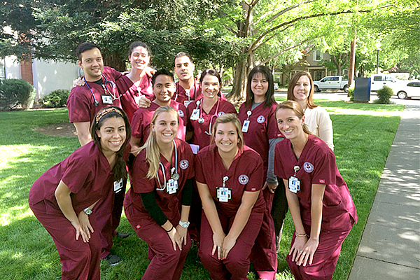 Ten nursing students stand in three rows outside wearing huge smiles.  Four women in front crouch slightly.  All ten students wear burgundy scrubs.  They stand with one woman not in hospital scrubs.