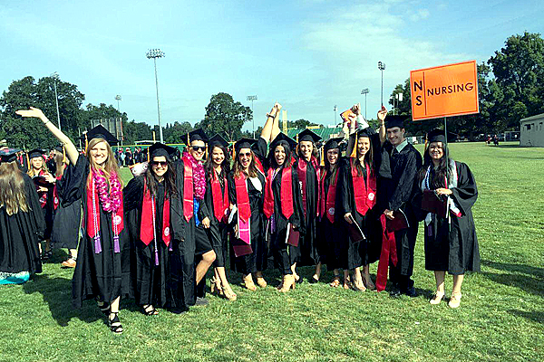 Eleven students in the Nursing program, dressed in black graduation gowns and caps, pose in a line for a photo.  Several students are adorned with cardinal red sashes around their shoulders while one student, on the far right, carries an orange sign that reads "NS| Nursing."