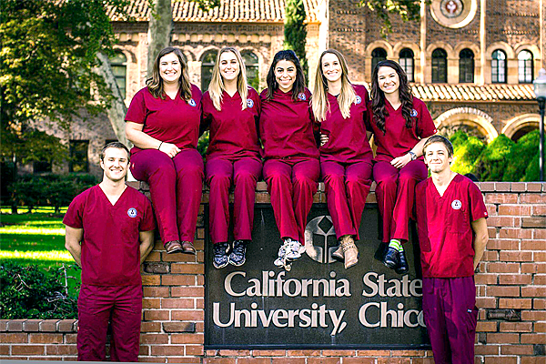 Five women sit on top of the California State University, Chico sign in front of Kendall Hall.  The women's feet dangles down in front of the sign encased by brick.  Two men stand on either side of the black metallic sign with silver lettering.