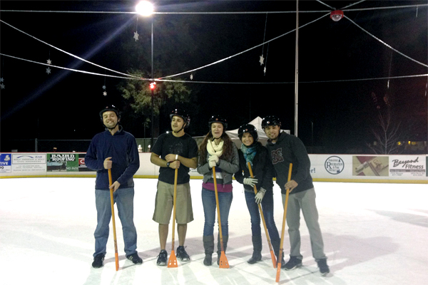 Five students wearing helmets, stand outside on a frozen rink.  One guy wears shorts as they all balance on sticks with small paddles that have holes in them.