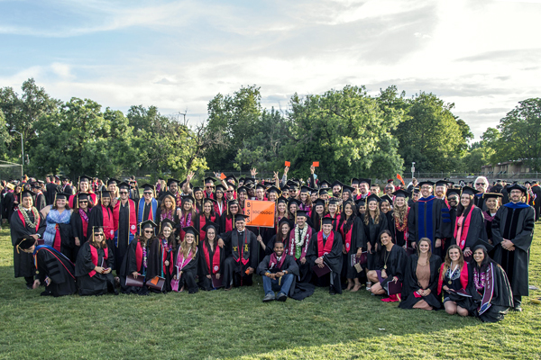 A group of sociology majors gathers for an afternoon photo.  They stand outside with partially gray skies and shadows creeping from right to left.  Almost all are dressed in graduate robes; some kneel, while some stand; some faculty pose on the right side of the photo.  