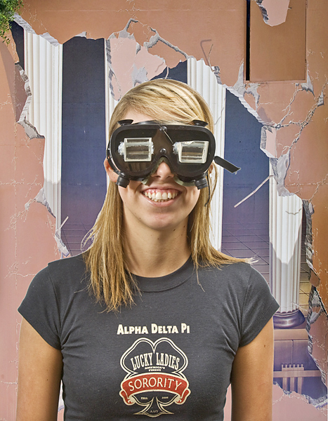 Smiling woman, superimposed over a background image of the John Pugh mural on the east side of Taylor Hall, wears strange pair of goggles.