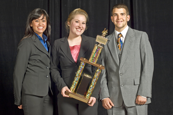 Three people stand in front of a black curtain. All 3 are dressed in business attire. From left to right: A woman with dark hair and mocha complexion stands with her arm behind her back, smiling, wearing a dark gray suit with royal blue collared shirt under the jacket. A woman with pulled back blonde hair stands center, wearing a dark gray suit with a pink blouse under the jacket while holding a large trophy. A man in a light gray suit, white shirt, and colorful tie stands to the right smiling.